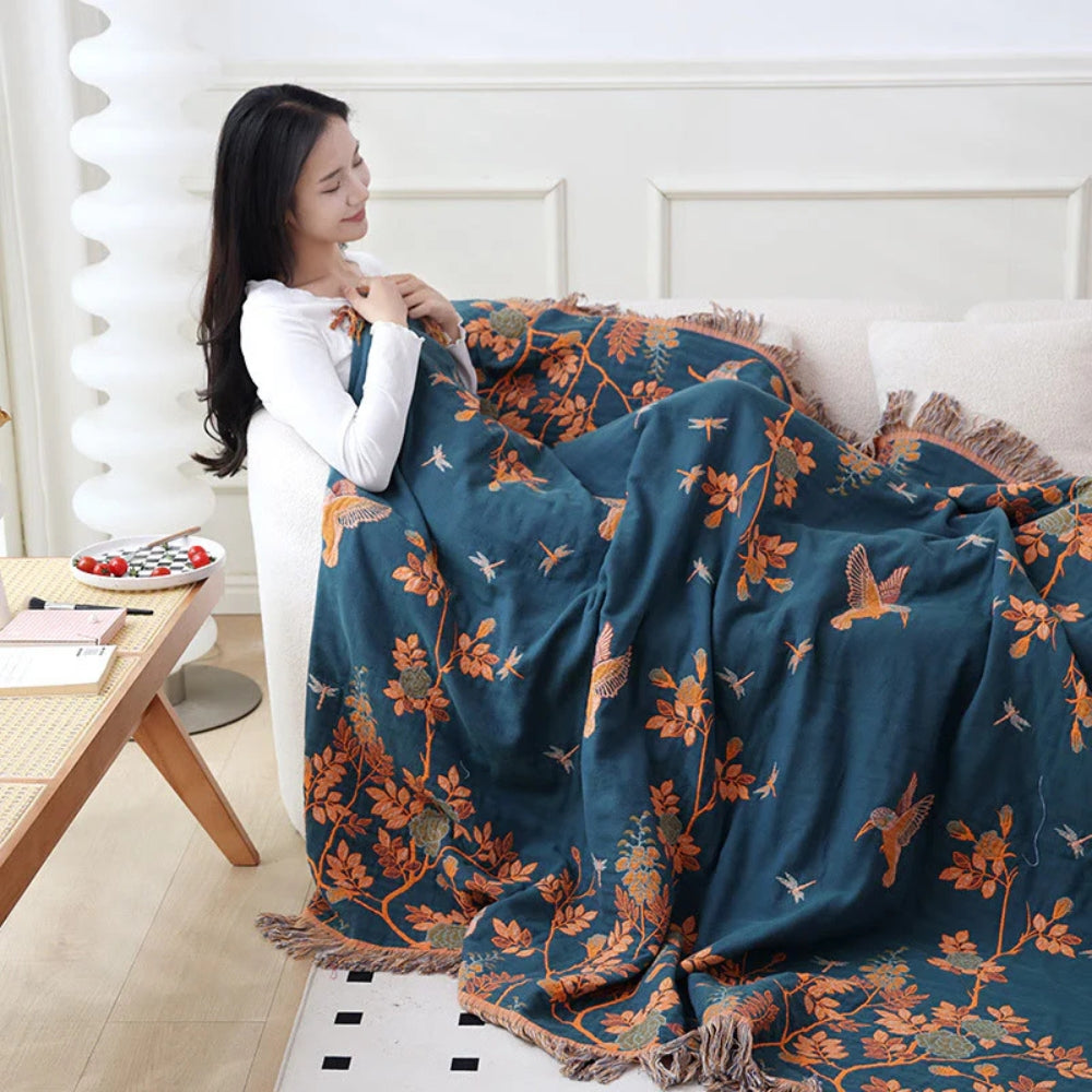 Woman wrapped in the dark blue bird and branch cotton blanket on a cream sofa, orange birds and branches and a fringed edge