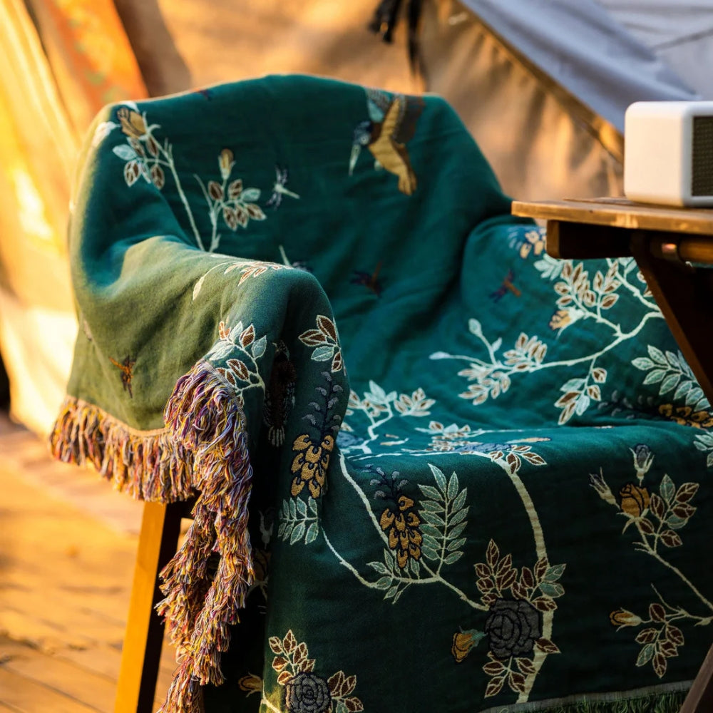 Teal bird and branch blanket draped over an armchair in low evening sun, a loose multicolored fringe hanging at the front