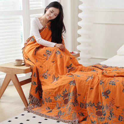 Orange bird and branch cotton blanket wrapped around a woman on a chair and spilling onto the bed, navy leaves and rust flowers