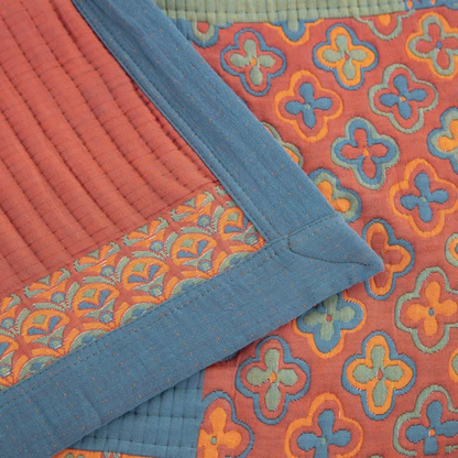 Close-up of the quilt corner: a mitered slate blue border with fine orange stitching, a rust channel-quilted block and woven flower and scallop patterns