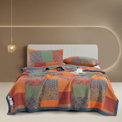 Patchwork cotton gauze quilt spread over a bed, plain orange, rust and sage blocks between small patterned blocks, with a slate blue border and two matching pillowcases