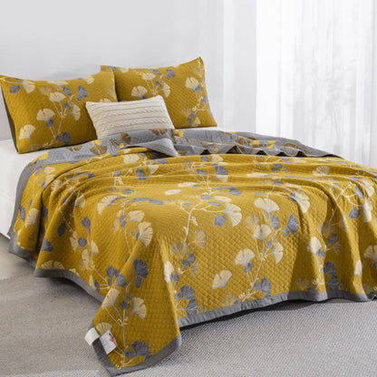 Mustard ginkgo quilt on a bed at an angle with cream and slate blue leaves, matching pillowcases and the gray reverse at the side