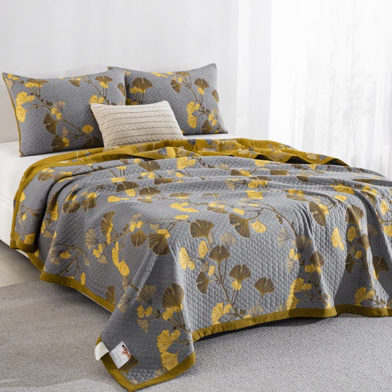 Gray ginkgo quilt on a bed at an angle with yellow and brown leaves, matching pillowcases and the mustard reverse and border