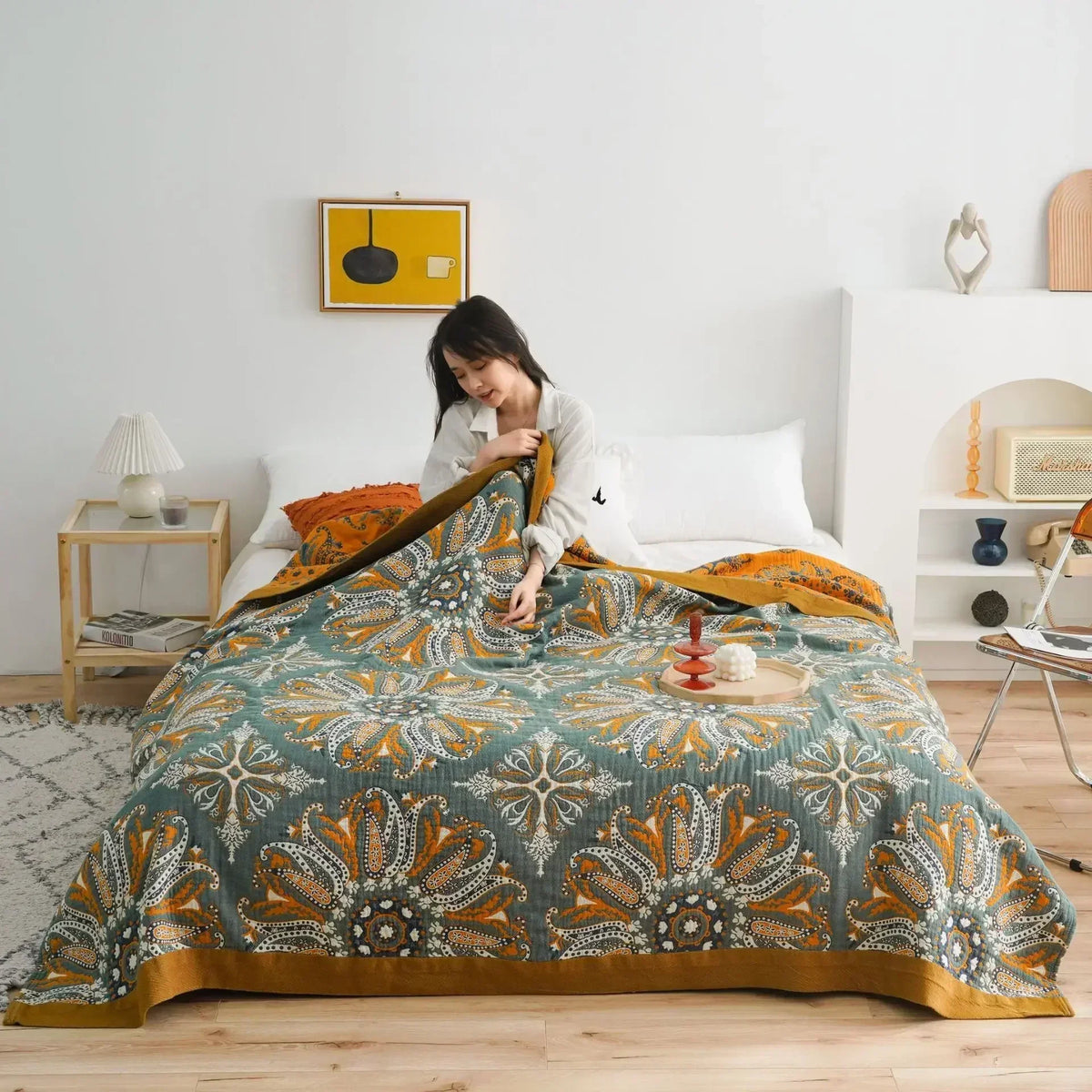 Dark teal paisley cotton quilt spread over a bed in a bright bedroom, a woman sitting up at the head holding one corner to show the orange reverse and mustard border