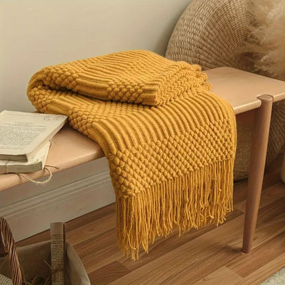 Mustard knit throw blanket folded on a wooden bench, zigzag bands of popcorn knots and slanted ribs with a tassel fringe hanging down