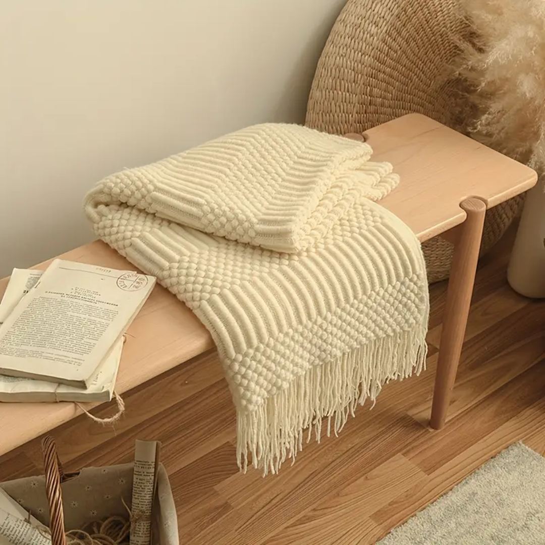 Cream knit throw blanket folded over a pale wooden bench, its raised popcorn and rib bands catching the light