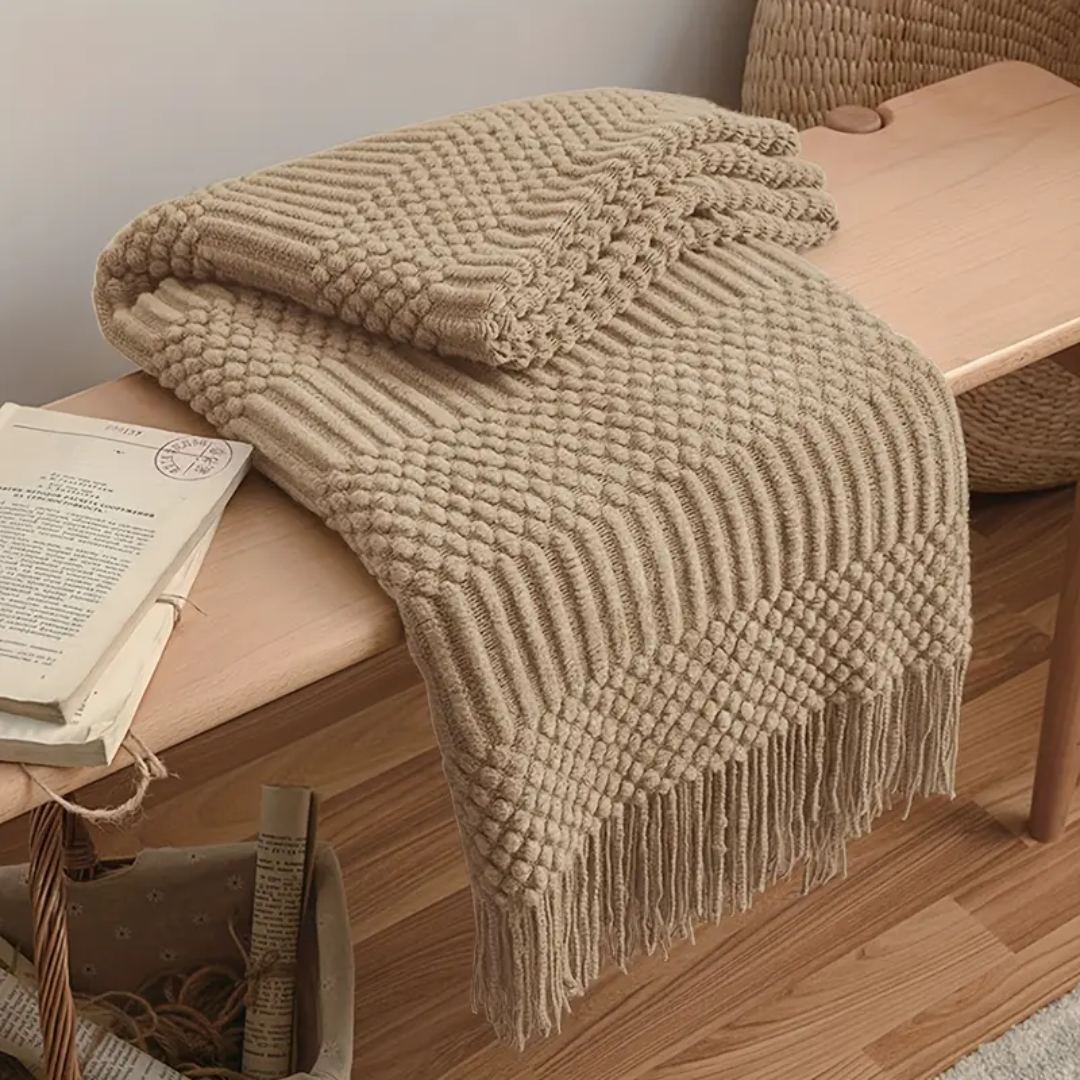 Tan knit throw blanket with tassel fringe folded over a wooden bench beside a rattan basket