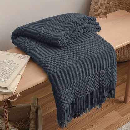 Charcoal gray knit throw blanket folded over a wooden bench, zigzag bands of knots and ribs ending in a tassel fringe