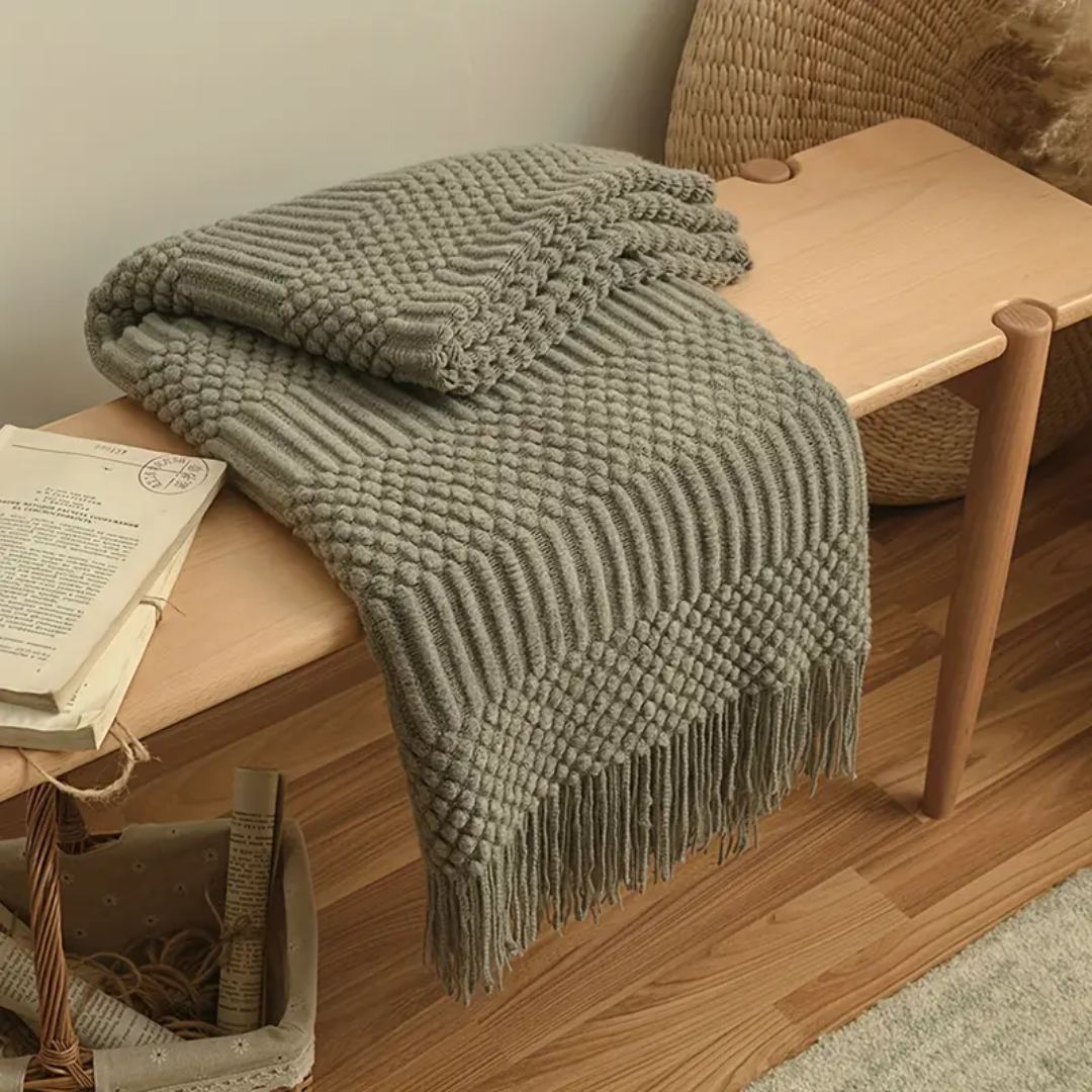 Olive gray-green knit throw blanket with tassels folded over a wooden bench