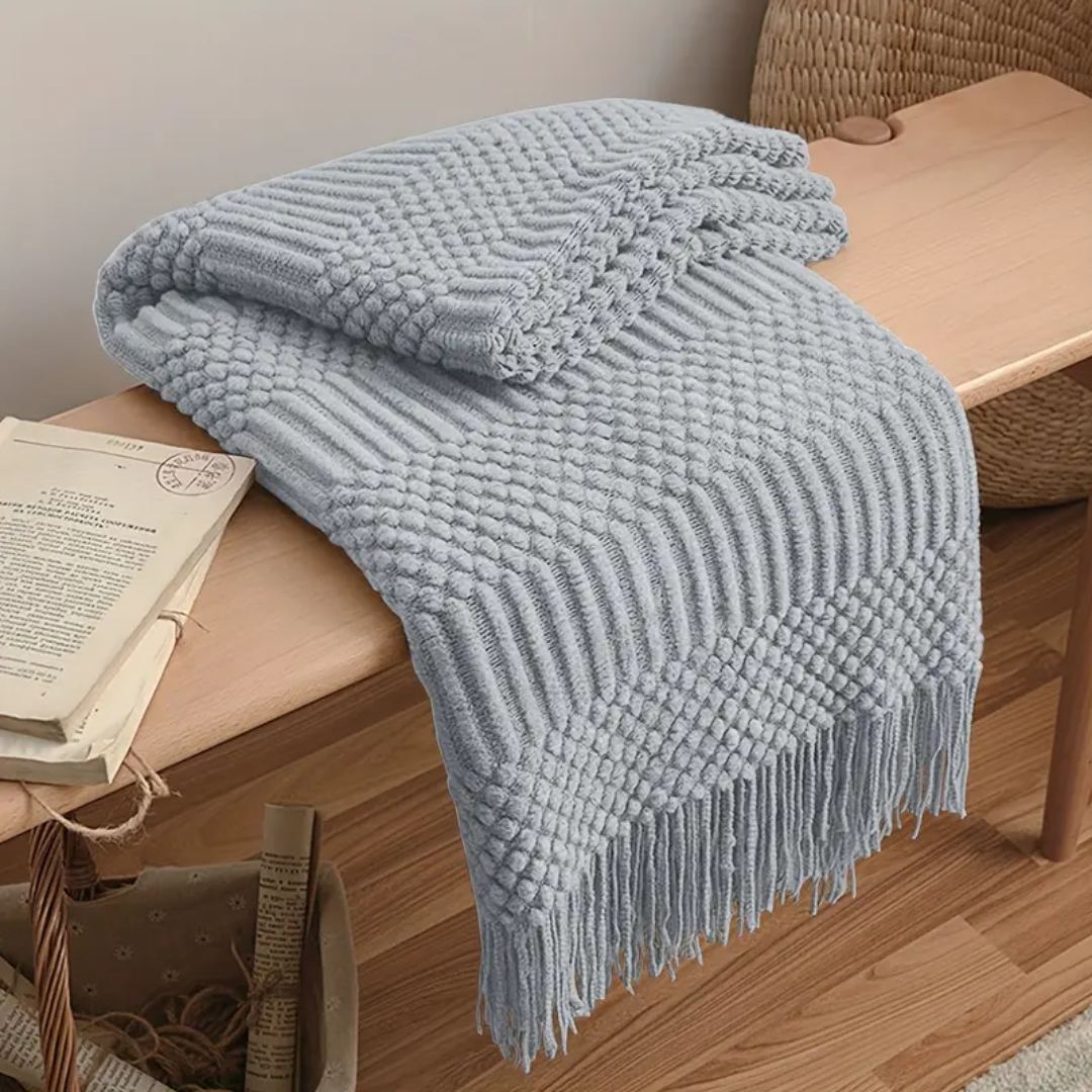 Pale gray textured knit throw blanket folded over a wooden bench, tassel fringe hanging toward the floor