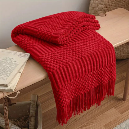 Red knit throw blanket folded over a wooden bench, the tassel fringe hanging below the seat
