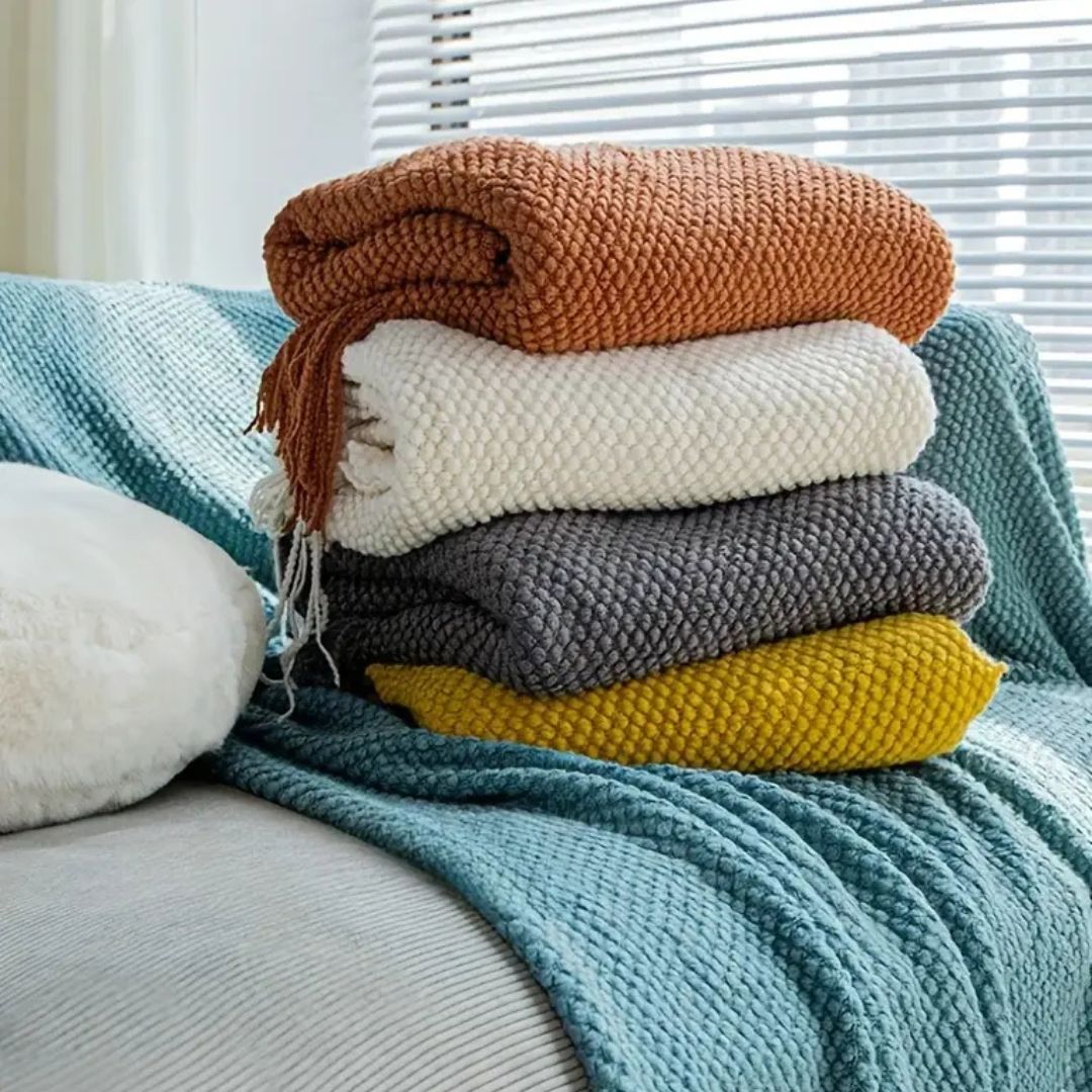 Stack of folded pebble knit throws with tassels on a sofa, rust, cream and mustard among them, the raised knit texture up close