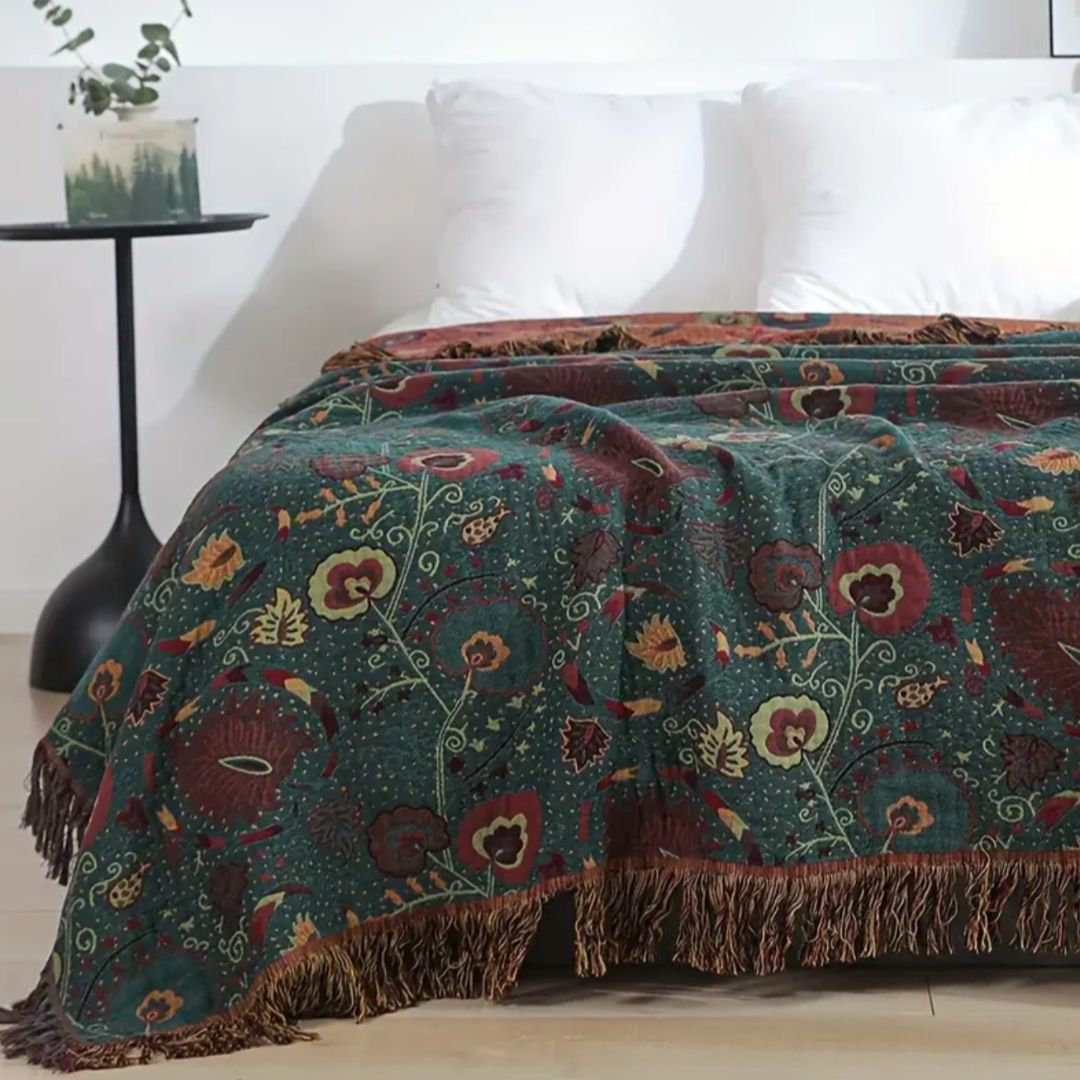 Close view of the reversible floral blanket over a bed, the dark teal-green face in front and the rust-orange side turned back at the head, with a loose fringe