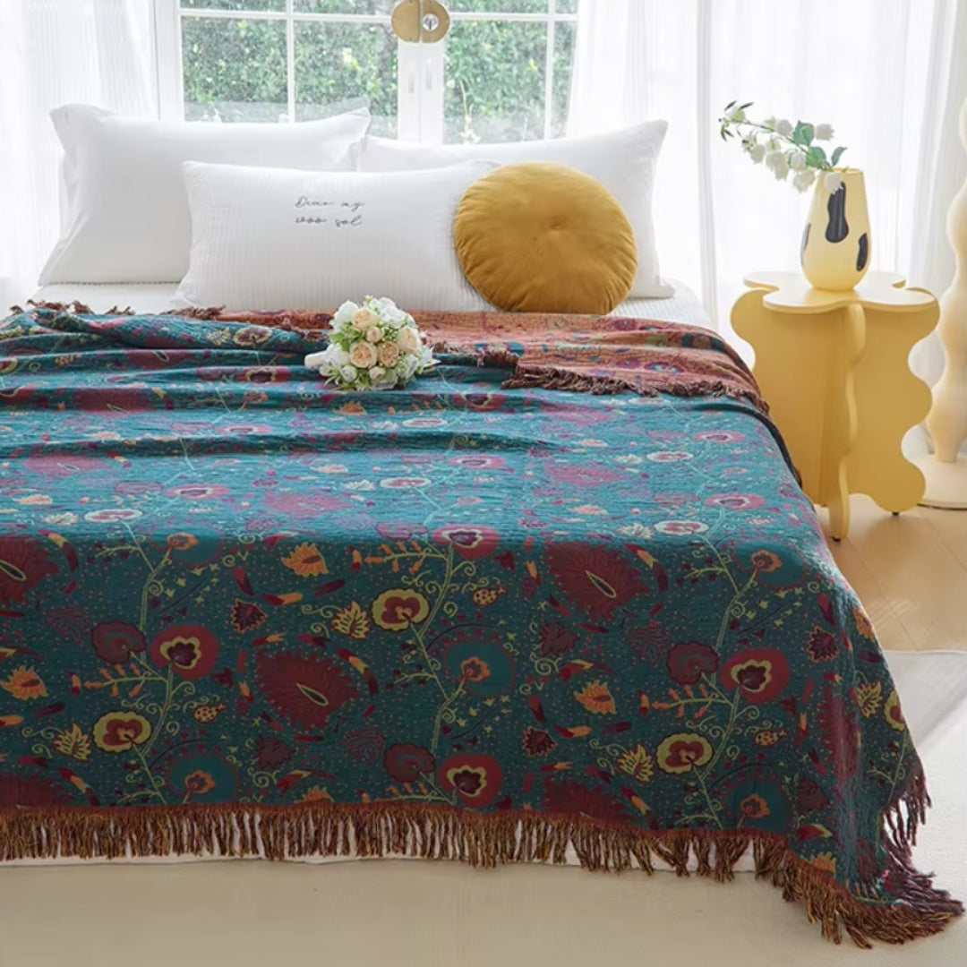 Floral cotton blanket on a bed beside a bright window, teal-green with burgundy flowers in front and the rust side folded back at the pillows