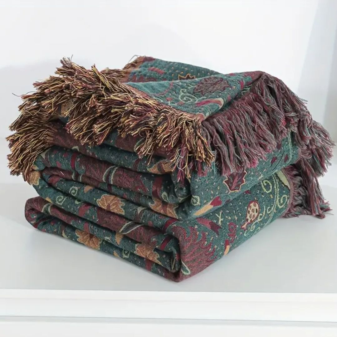 The floral blanket folded on a white shelf, its teal-green weave and burgundy, rust and gold fringe in front