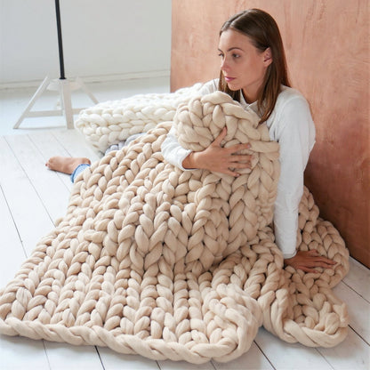 Woman sitting on white-painted floorboards wrapped in a beige chunky knit throw, with a white one bunched behind her
