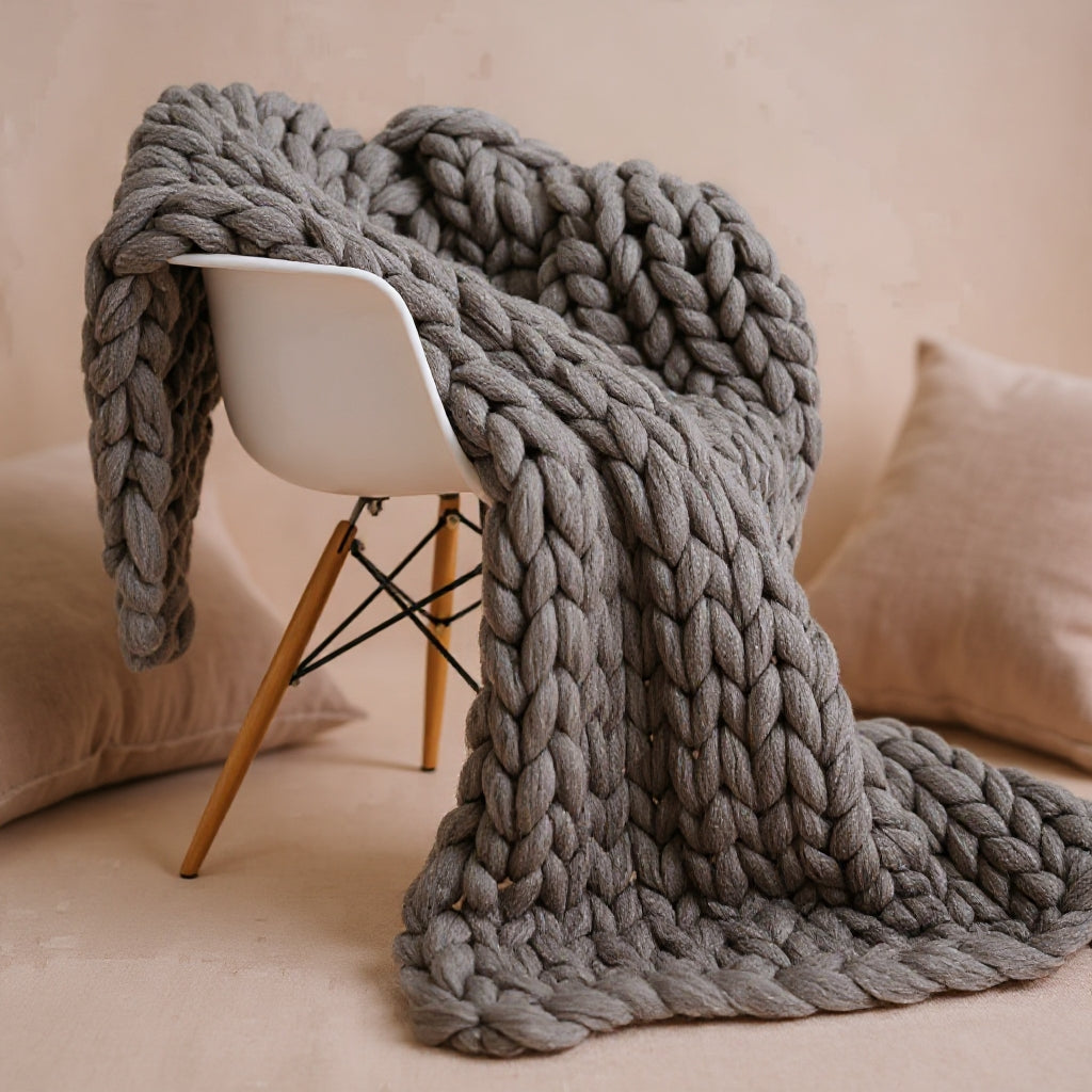 Gray chunky knit throw draped over a white shell chair, the loops running in thick chains down to the braided edge