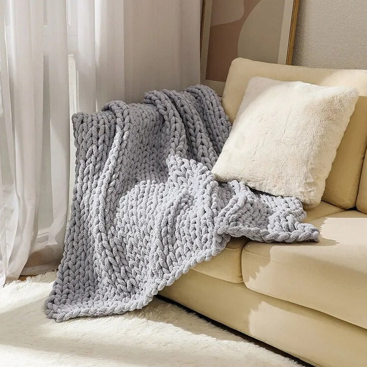 Silver chunky knit throw spread over the corner of a sofa beside a cream faux fur cushion, its chained edge resting on the rug