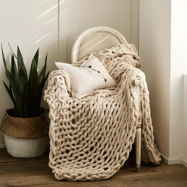 Beige chunky chenille throw draped over a white wooden chair with a linen cushion, beside a snake plant in a woven basket