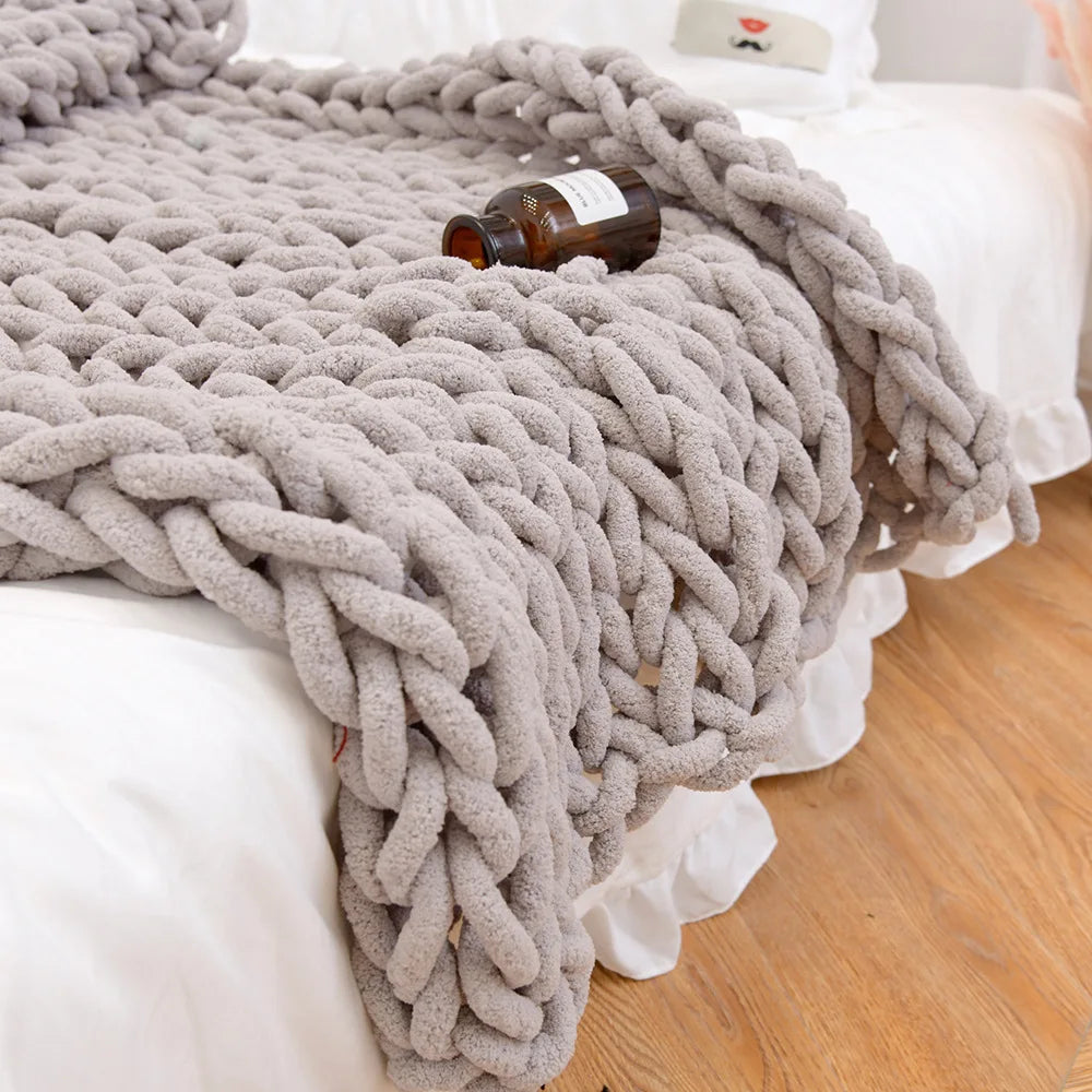 Taupe chunky chenille knit throw falling over the end of a white bed onto a wood floor, a small amber bottle resting on its loops