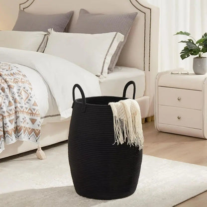 A black rope laundry hamper with a cream throw hanging over the rim, beside a bed.