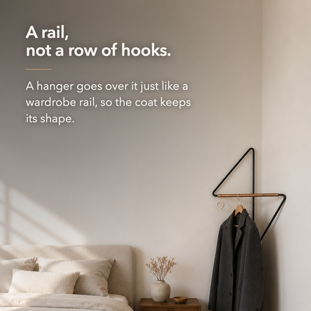Bedroom corner with the coat rack holding a dark coat on a hanger, and overlaid text reading: A rail, not a row of hooks. A hanger goes over it just like a wardrobe rail, so the coat keeps its shape.