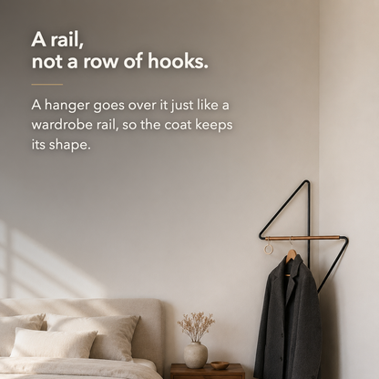 Bedroom corner with the coat rack holding a dark coat on a hanger, and overlaid text reading: A rail, not a row of hooks. A hanger goes over it just like a wardrobe rail, so the coat keeps its shape.