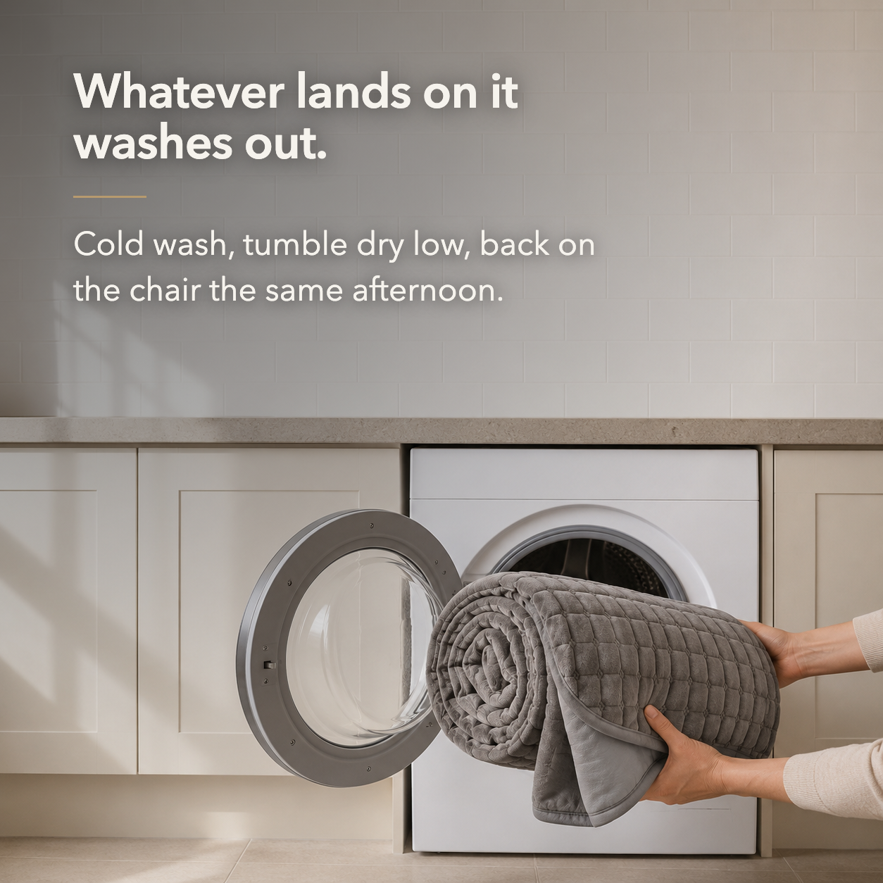 Hands feeding the rolled gray quilted cover into a front loading washing machine, with the words Whatever lands on it washes out