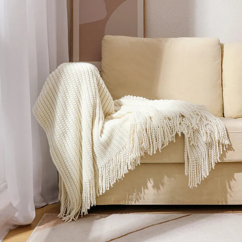 A cream bobble knit throw blanket with fringed ends draped over the arm of a sofa.