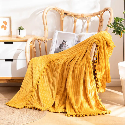 A mustard flannel throw with raised stripes and mustard pom-poms over a rattan armchair