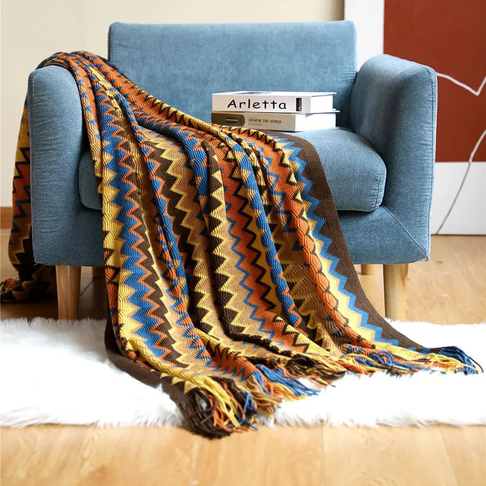 An orange chevron throw with mustard, blue and brown bands folded over a blue armchair, its tassels on a white rug.