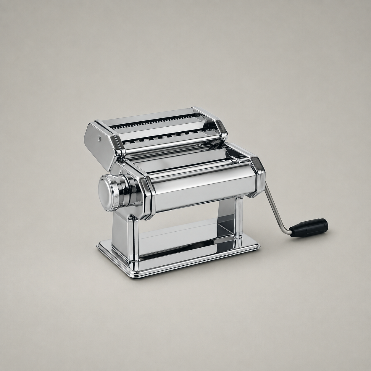 Stainless steel manual pasta maker with crank handle, on a plain warm grey studio background