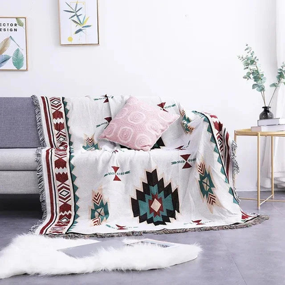 A white jacquard throw with one large black and teal medallion and burgundy borders, over a gray couch