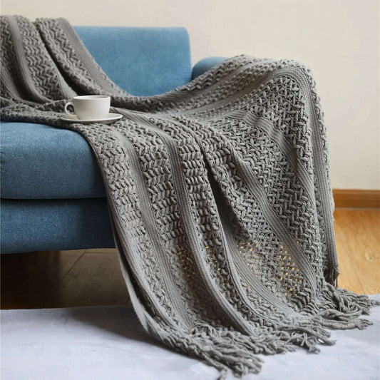 Gray crochet knit throw draped over a blue sofa and onto the floor, showing stripes of raised V stitches, open lattice and a knotted tassel fringe