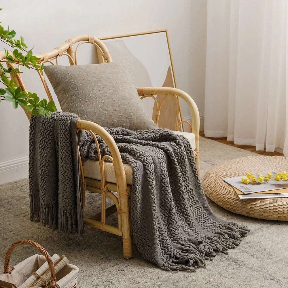 Gray knit throw with tassels laid over a rattan armchair beside a woven floor cushion in a bright room