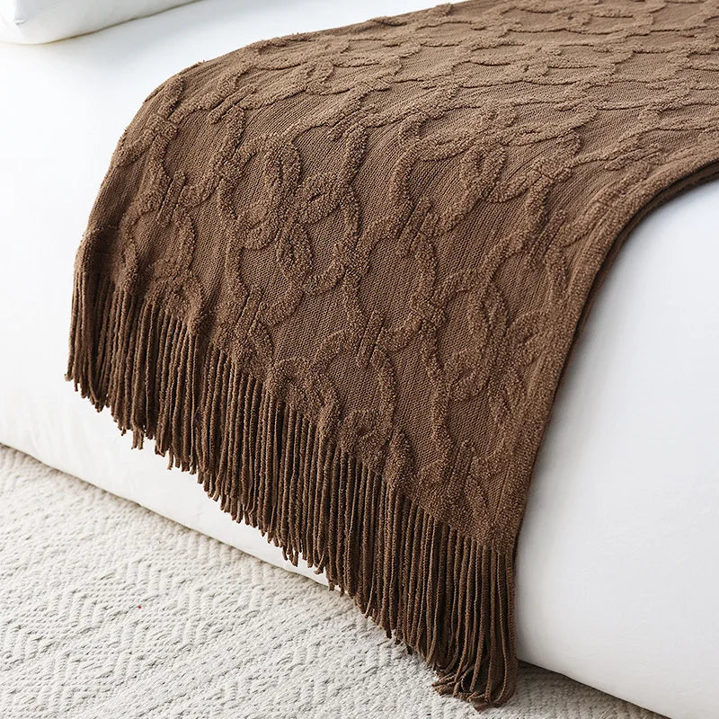 Brown jacquard knit throw draped over the end of a white bed, its raised ring pattern catching the light and a long tassel fringe hanging to the rug