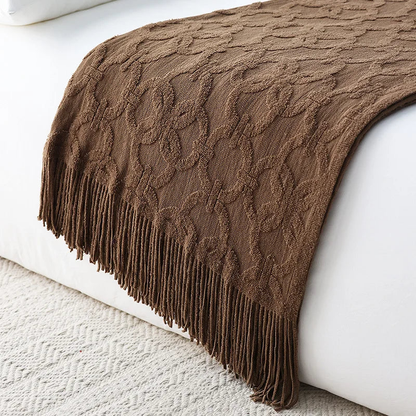 Brown jacquard knit throw draped over the end of a white bed, its raised ring pattern catching the light and a long tassel fringe hanging to the rug