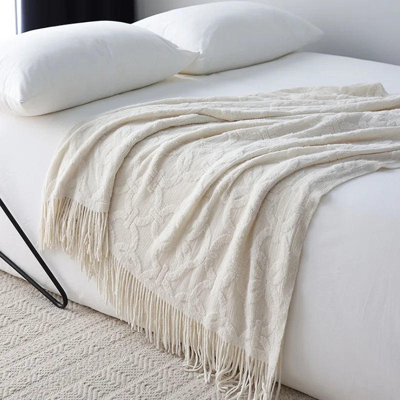 White knit throw with a raised ring and crossbar pattern draped across a white bed, the tassel fringe falling over the side