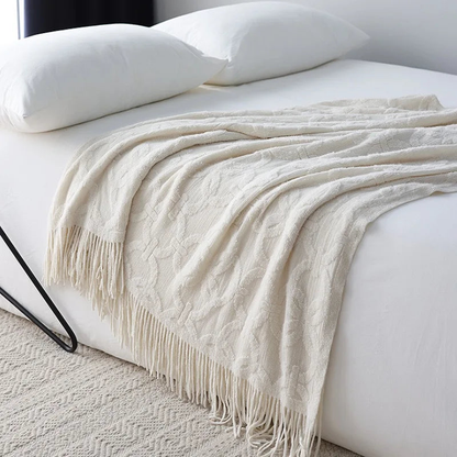 White knit throw with a raised ring and crossbar pattern draped across a white bed, the tassel fringe falling over the side