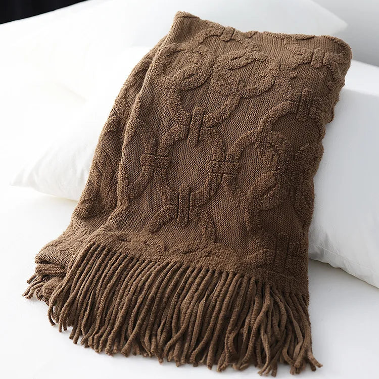 Brown jacquard knit throw folded over white pillows, showing the raised looped rings and the long fringe