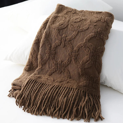 Brown jacquard knit throw folded over white pillows, showing the raised looped rings and the long fringe