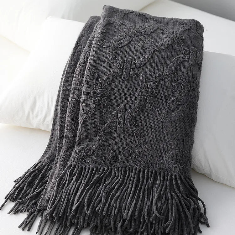 Charcoal knit throw folded over white pillows, its interlocking ring pattern in raised loops and a long tassel fringe