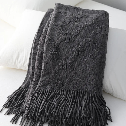 Charcoal knit throw folded over white pillows, its interlocking ring pattern in raised loops and a long tassel fringe
