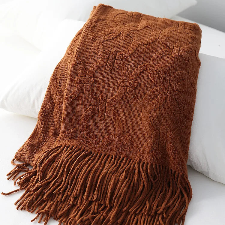 Rust jacquard knit throw folded over white pillows, the raised ring lattice and tassel fringe in burnt orange