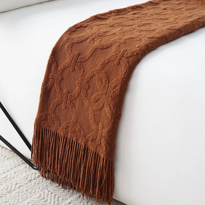 Rust knit throw laid like a runner over the edge of a white bed, its raised ring pattern and tassel fringe in burnt orange