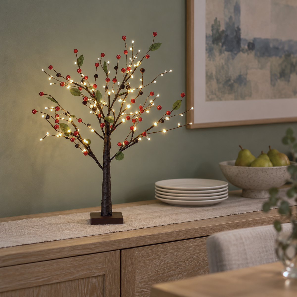 Red berry tabletop tree lit on an oak dining sideboard with a linen runner, stacked plates and a bowl of pears against a sage green wall