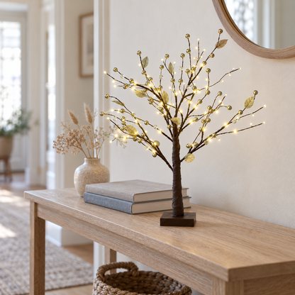 Gold tabletop twig tree lit on an oak hallway console table beside books and a vase of dried grasses, under a round mirror in daylight