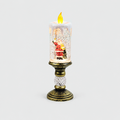 Christmas snow globe candle lit on a light gray background, a red-suited Santa with a teddy bear and a bell inside the glittering clear column, under a white drip cap and an LED flame, on an antique gold-look pedestal