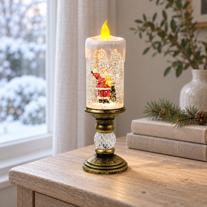 Lit snow globe candle with Santa holding a bell and a teddy bear inside, on an oak side table by a window with snowy trees outside, beside a stack of linen books and a pine sprig
