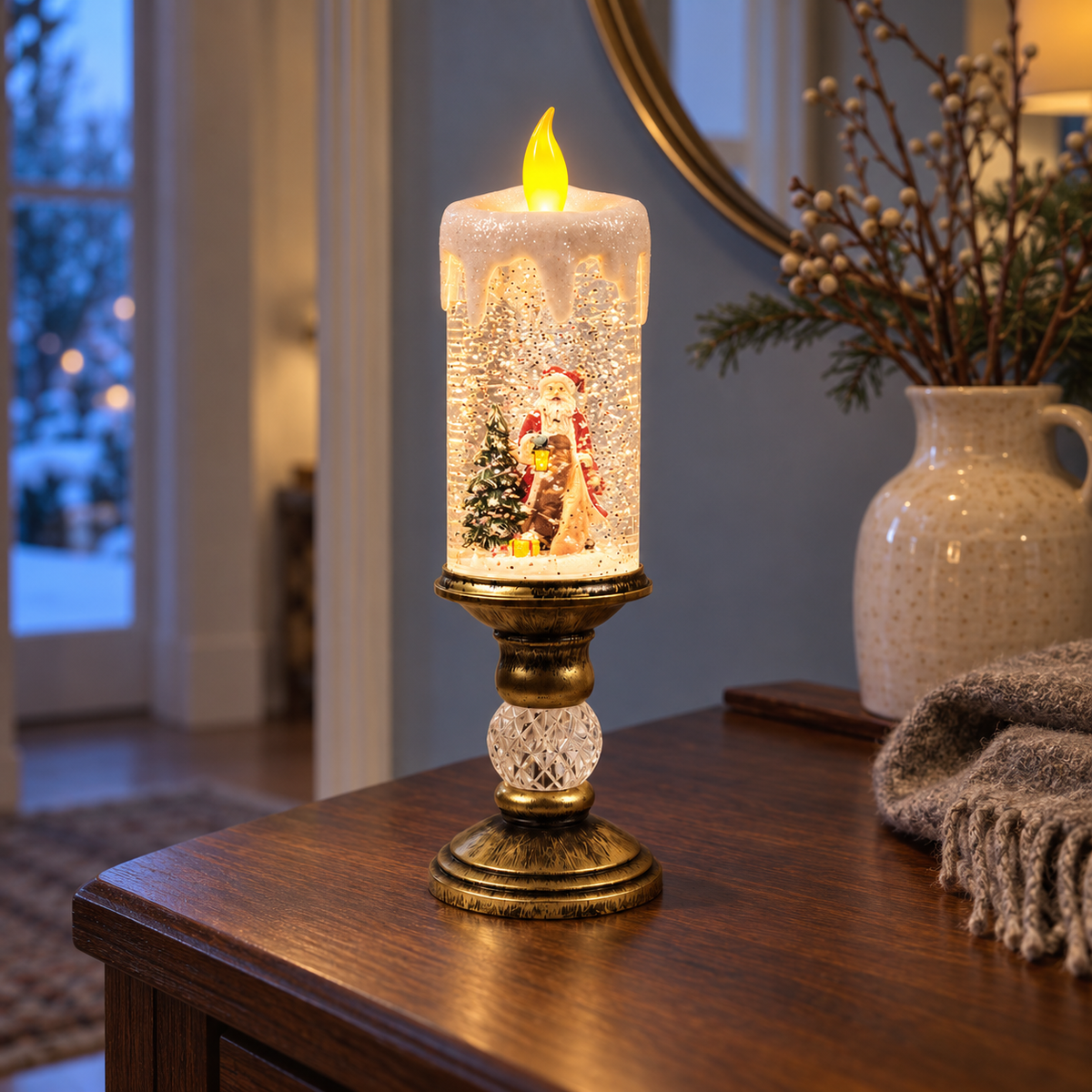 Lit snow globe candle with Santa holding a lantern beside a snowy tree, on a dark wood hallway console at dusk, next to a cream jug of berry branches and a knitted throw